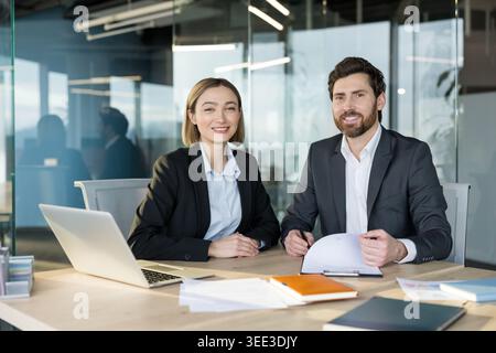 Business professionals smiling while sitting at a desk together, working on projects and discussing ideas in a contemporary corporate office environment Stock Photo