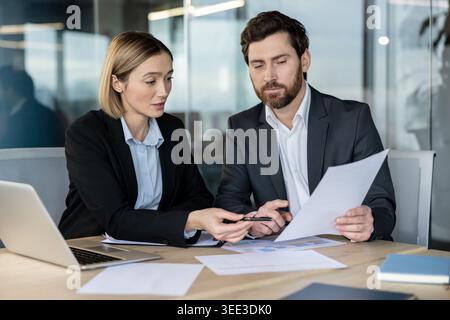 Two colleagues in a modern office review financial charts and paperwork, pointing at a laptop and documents as they collaborate on strategy and planning during a meeting Stock Photo