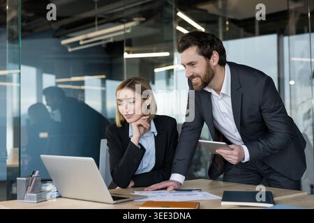 Two business professionals in formal wear collaborating on a project, analyzing data on a laptop and tablet, working together in a modern office environment Stock Photo