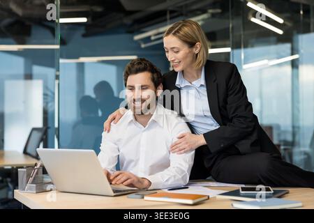 Two smiling adult coworkers, a man and a woman, actively collaborating and finding success while viewing information on a laptop in a contemporary office environment Stock Photo