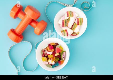 Creative composition made of orange dumbbells, colorful gummy candies and measuring tape on pastel blue background. Fitness, healthy lifestyle idea. Stock Photo