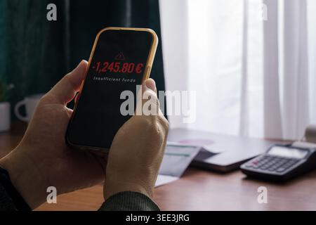 Adult man holding smartphone with banking app showing insufficient funds warning and negative balance, indoor home setting with calculator and documen Stock Photo