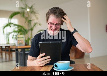 Scandinavian man sitting at a wooden table in a bright cafe, scratching his head with a stressed, frustrated expression while looking at a digital tab Stock Photo