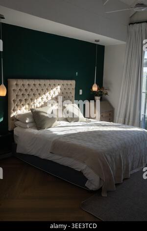 Double bed with tufted beige headboard sitting in center of bedroom, catching sun through curtains Stock Photo