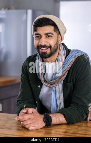 Indian man in thirties leaning on wooden countertop in kitchen wearing beanie and scarf Stock Photo