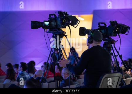Professional camera operator filming corporate conference, live video production with crowd, tripod, headset, purple blue lighting. Stock Photo