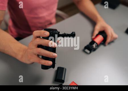 Photographer hands inserting red rechargeable battery pack into camera battery grip on gray table, prepping professional gear for shooting in dark stu Stock Photo