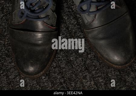 Overhead view of dark worn leather boots with blue laces on a textured grey carpet grounding concept for urban fashion and style clothing Stock Photo