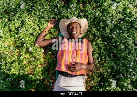 top view of happy woman lying on green grass blooming white flowers spring forest wearing straw hat striped top looking up smiling enjoying nature Stock Photo