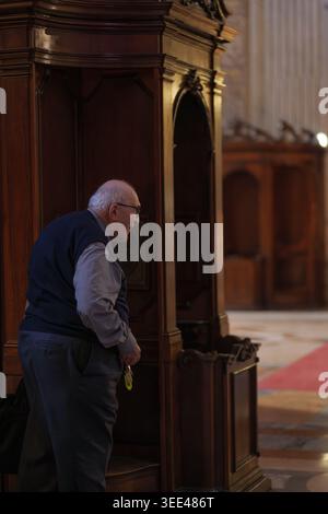 An older man, with glasses, stands next to an ornate wooden confessional booth inside a church in Bologna, Italy. The setting is dimly lit, highlighti Stock Photo