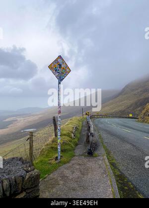 Sticker-covered road warning sign along the winding descent of Conor Pass, Dingle Peninsula, with valley and lake views below, Kerry, Ireland Stock Photo