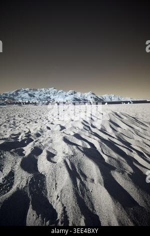 An infrared-style photograph capturing the texture and forms of a sandy beach in the foreground with contrasting dark shadows. In the background, what Stock Photo