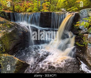 A small waterfall cascades over rocks surrounded by lush greenery. The flow of water creates a calming atmosphere, enhanced by the autumn colors of th Stock Photo