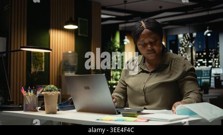Businesswoman does critical thinking and proofreading documents before transcribing information on notebook, standing in the office at night time. Documenting important ideas. Camera A. Stock Photo