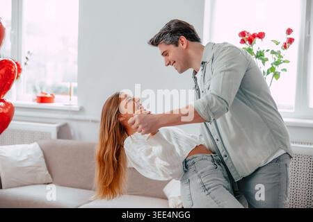 Happy young couple dancing playfully at home, smiling romantic partners holding hands during love celebration in bright modern living room Stock Photo