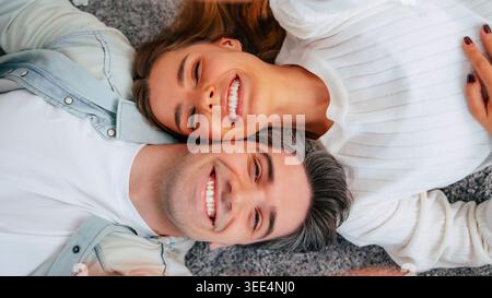 Happy young couple lying on carpet at home, smiling romantic partners laughing together in cozy modern living room Stock Photo