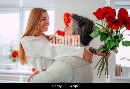 Happy young woman being lifted by boyfriend after receiving red roses at home, romantic couple celebrating love in bright modern living room Stock Photo
