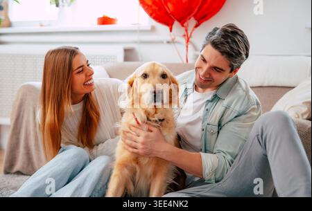 Happy young couple relaxing with their dog at home, smiling pet owners bonding with golden dog in cozy modern living room Stock Photo