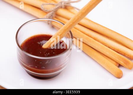 a breadstick dipped with ketchup dipping sauce in transparent cup with pack of breadsticks in background in white dish Stock Photo