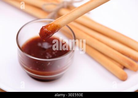 a breadstick dipped with ketchup dipping sauce in transparent cup with pack of breadsticks in background in white dish Stock Photo
