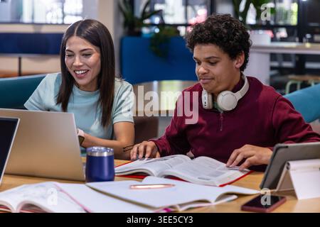 Diverse students collaborating at wooden table in study lounge using laptops, textbooks, headphones Stock Photo