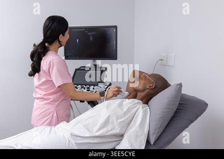 Diverse female clinician in scrubs, patient lying on exam bed having ultrasound using probe, screen Stock Photo