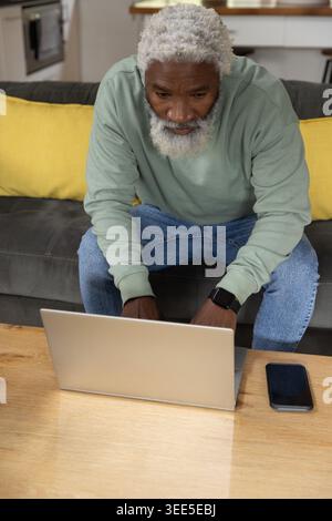 Senior African American man leaning forward on gray sofa using silver laptop with smartphone nearby Stock Photo
