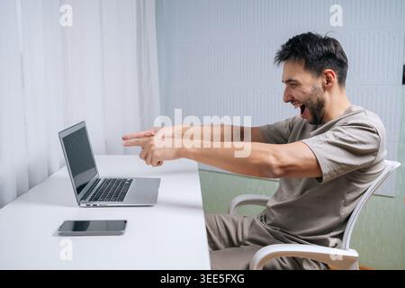 Side view of man actively working on laptop, reacting with extreme excitement, joy, and triumphant shout, pointing with both hands at screen, celebrat Stock Photo