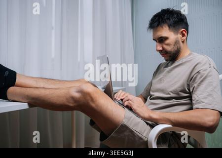 Relaxed freelancer male enjoying comfortable remote work at home, typing on laptop with feet casually resting on desk, embodying flexibility and moder Stock Photo