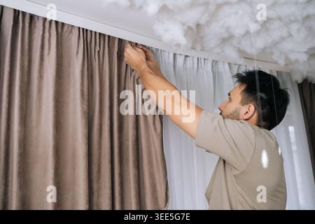 Low-angle view of man installing curtains on white rail, attaching fabric hooks to refresh a living room window during a home renovation, surrounded b Stock Photo