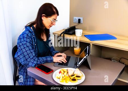 Woman working remotely during breakfast on laptop in hotel room Stock Photo