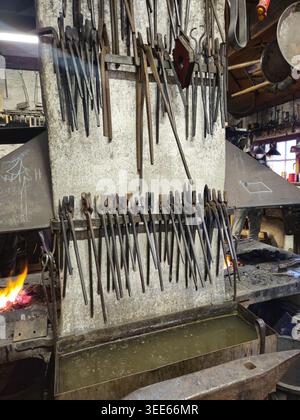 Rustic blacksmith workshop interior with rows of hanging tongs and pliers on a soot-stained wall, glowing coals and an anvil beside a quenching trough Stock Photo