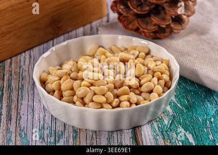 A close up shot of a white ceramic bowl filled with shelled pine nuts. It sits on a rustic teal brown wooden table with a pine cone linen cloth in the Stock Photo