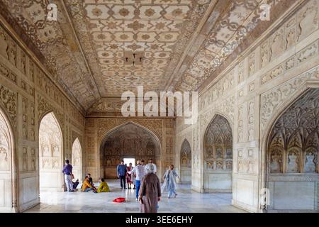 Ornate white marble interior of a historic Mughal palace hall featuring intricate carvings and patterned ceiling with tourists in Agra Fort India Stock Photo