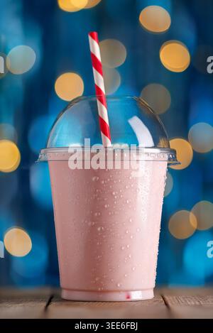Chilled strawberry milkshake in disposable plastic cup with condensation and red and white striped straw on wooden table Stock Photo