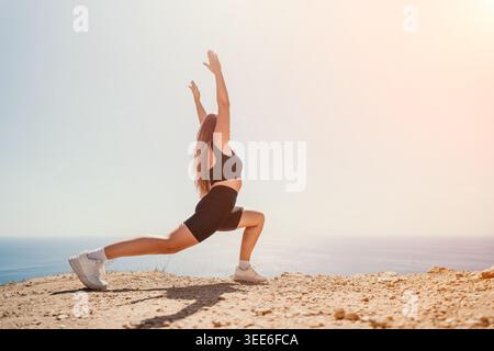 Yoga stretching fitness woman in black sportswear practicing a lunge pose on a sunny cliff overlooking the ocean with copy space Stock Photo