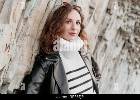 Woman leather jacket rock Portrait of stylish woman in striped turtleneck and jacket leaning against textured cliff Stock Photo