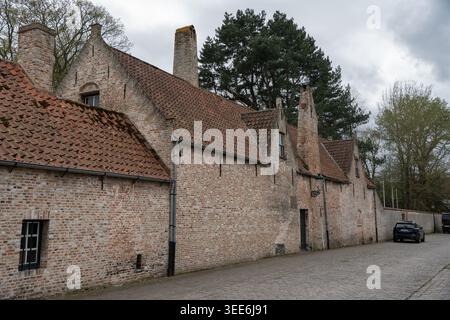 Bruges, Belgium - April 4, 2026: Gezellehuis brick wall, red tiled roof, chimney, small window, cobblestone street, parked car, and trees appear under Stock Photo