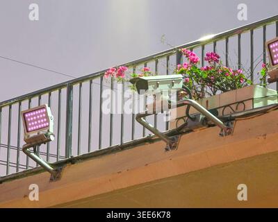Security camera and floodlight installed on metal railing of outdoor balcony with bright pink flowers providing sense of safety and calm observation a Stock Photo
