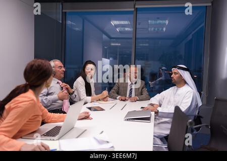 A diverse group of Middle Eastern and international executives in a serious business meeting inside a modern glass-walled office, using laptops and notebooks to discuss strategy, negotiation and corporate planning. Stock Photo