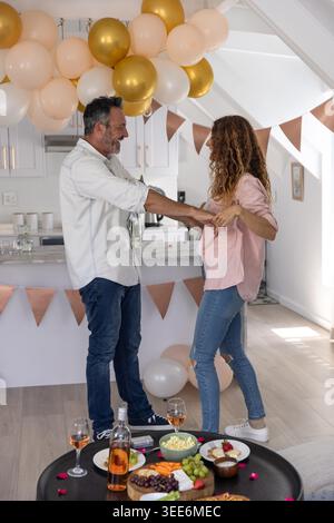 Couple clinking rose glasses in kitchen, wearing shirts and jeans with charcuterie and balloons. Celebration, romance, home, decor, balloon, garland, sunlit Stock Photo