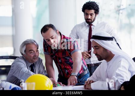 A multicultural business team reviews construction plans together, with engineers and managers collaborating over blueprints and a safety helmet, focusing on project planning, teamwork and decision-making. Stock Photo