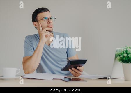 Serious pensive male wears round glasses calculates expanses uses generic laptop computer and calculator, surrounded with mug of coffee and documentation, sits at coworking space. Business concept Stock Photo