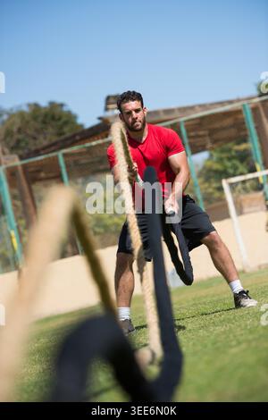 Middle Eastern Arab man performing an intense outdoor CrossFit battle rope exercise for strength, conditioning and athletic training in an open-air fitness area, showing focus, power and endurance. Stock Photo