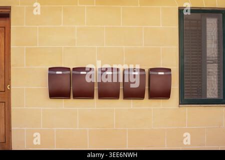 Several identical brown mailboxes on a yellow limestone wall between a door and a window. Concept of communication, postal service and house exterior. Stock Photo