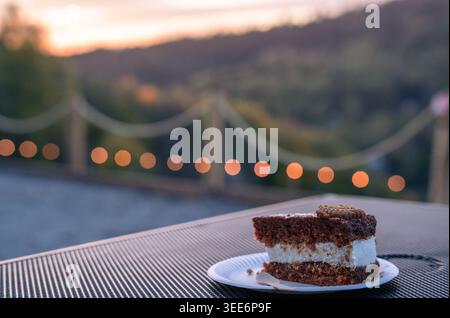 A delicious piece of chocolate layer cake with white cream served on a small plate outdoors Stock Photo