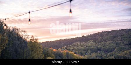 A decorative string of glowing light bulbs hanging outdoors against a blurry sunset forest and hills background Stock Photo