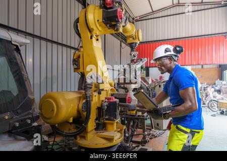 Adult African engineer uses a laptop to program an industrial robot in a workshop for automation, maintenance, and engineering concepts in Asia. Stock Photo