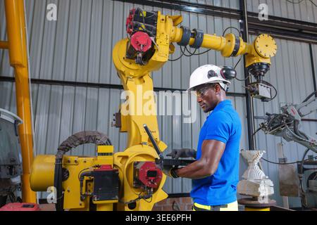 Adult African engineer uses a laptop to program an industrial robot in a workshop for automation, maintenance, and engineering concepts in Asia. Stock Photo