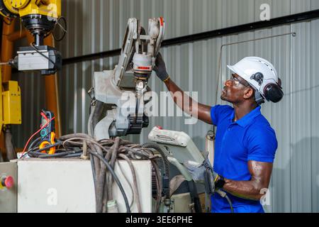 Adult African engineer uses a laptop to program an industrial robot in a workshop for automation, maintenance, and engineering concepts in Asia. Stock Photo